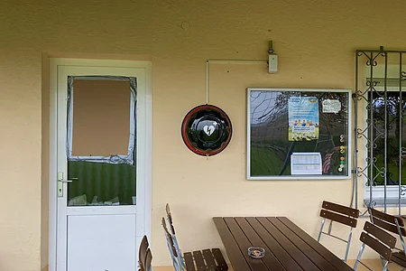 Außenwand mit weißer Tür, defektem Fenster, Defibrillator und Holztisch mit Stühlen.