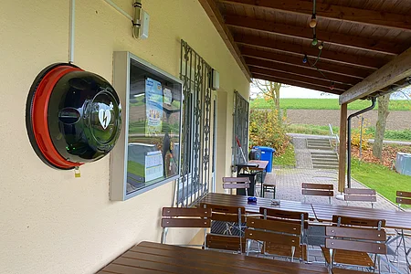 Überdachte Terrasse mit Holztischen, Stühlen und einem an der Wand montierten Defibrillatorkasten.