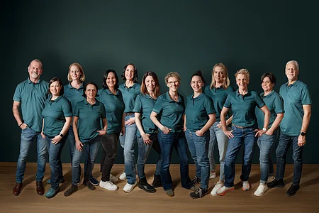 Gruppenfoto von 14 Personen in blauen Poloshirts und Jeans vor dunklem Hintergrund stehend.