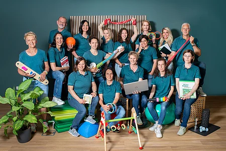 Gruppenfoto von 16 Personen in blauen Poloshirts mit verschiedenen Therapie- und Trainingsgeräten vor grünem Hintergrund.