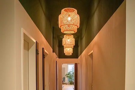 Langer Flur mit Holzboden, weißen Türen und drei hängenden Lampen, am Ende ein Raum mit Pflanze.