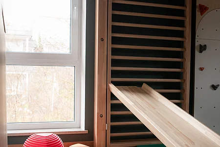 Raum mit Holzrutsche, Kletterwand und bunten halbrunden Balanciersteinen auf hellem Boden.