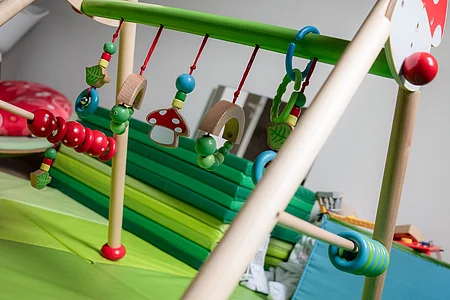Holz-Babyspieltrapez mit bunten hängenden Spielzeugen auf grünem Spielteppich in einem Raum.