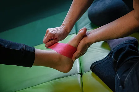 Person wickelt roten Tapeverband um den Knöchel einer anderen Person auf grünem Untergrund.