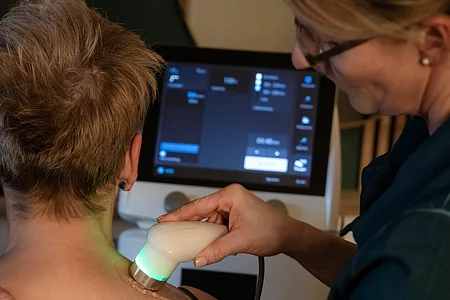 Frau erhält Ultraschallbehandlung am oberen Rücken durch medizinische Fachkraft mit Gerät und Monitor.