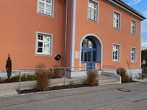 AED - Schulhaus Dünzling Eingang eines orangefarbenen Gebäudes mit blauer Tür, mehreren Fenstern und Rampe mit Geländer.