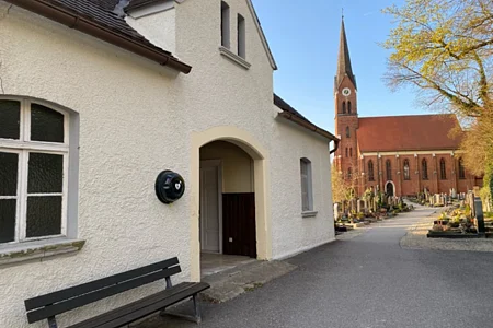 Weißes Gebäude mit Bank davor und Kirche mit Friedhof im Hintergrund bei klarem Himmel