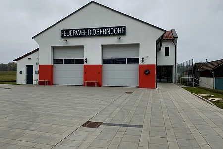 Feuerwehrhaus Oberndorf mit zwei geschlossenen Toren und rotem Sockel bei bewölktem Himmel.