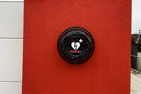 Defibrillator in schwarzem Gehäuse an roter Wand montiert, links weiße Wandpaneele sichtbar.