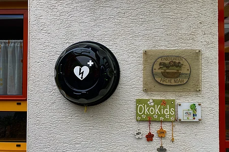 Defibrillator an weißer Wand neben Schildern mit Kindergarten- und Ökokids-Schriftzügen und bunten Anhängern.