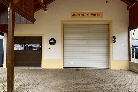 Gebäude mit zwei geschlossenen Toren und Schild „Feuerwehr - Gerätehaus“ über dem größeren Tor.