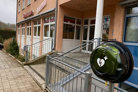 Außenansicht eines Gebäudes mit Sparkasse-Schriftzug, Eingangstreppe und Defibrillator an Geländer.