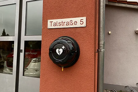 Defibrillator an roter Hauswand unter Straßenschild Talstraße 5 neben grauer Tür und Regenrinne