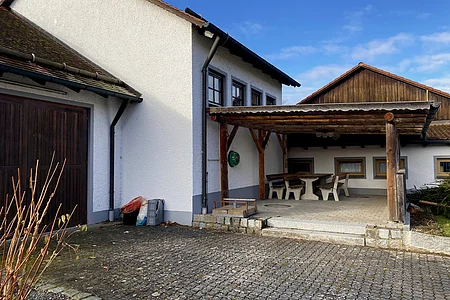 Außenbereich eines Hauses mit überdachter Sitzgruppe aus Holz und gepflastertem Hof bei klarem Himmel.