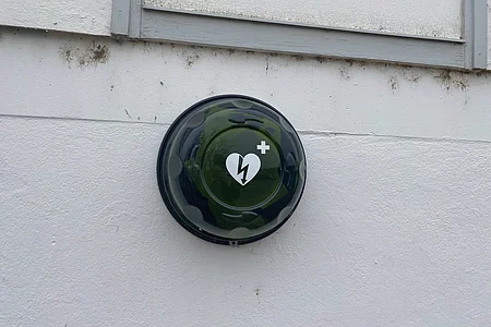 Grünes rundes Gehäuse mit Herzsymbol und Blitz, befestigt an weißer Außenwand, Defibrillator-Schild.
