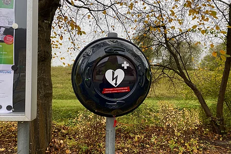 Außen an einem Baum befestigtes schwarzes Gehäuse für einen Defibrillator im Herbst mit gelben Blättern.