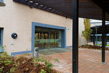 Eingang eines Gebäudes mit Glas-Schiebetür, davor Pflanzbeet und überdachter Weg mit Holzsäulen.