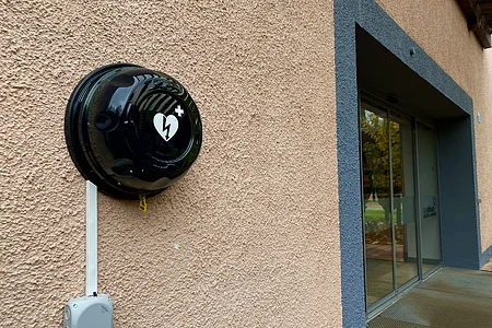 Defibrillator in schwarzem Gehäuse an einer Außenwand neben einem Eingang mit Glastür.
