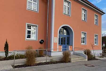 Eingang eines orangefarbenen Gebäudes mit blauer Tür, mehreren Fenstern und Rampe mit Geländer.