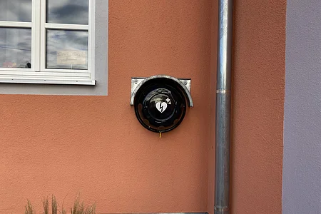 Defibrillator-Halterung an einer orangefarbenen Hauswand neben einem Fenster und einem Fallrohr.