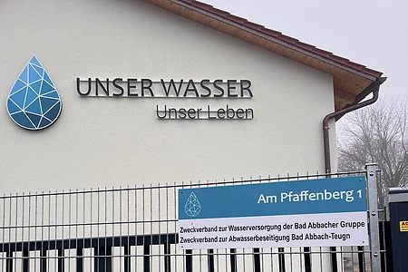 Gebäudewand mit Schriftzug „Unser Wasser Unser Leben“ und Zaun mit Schild „Am Pfaffenberg 1“ und Defibrillatorkasten.