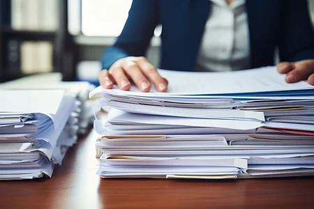Zwei große Stapel mit Papierdokumenten auf einem Tisch, eine Person sortiert die Unterlagen.