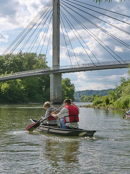 Personen in Kanus mit Schwimmwesten paddeln auf einem Fluss unter einer Fußgängerbrücke.