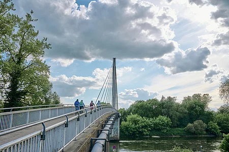 Eine Brücke mit Geländern, auf der Menschen mit Fahrrädern fahren. Darunter fließt ein Fluss, umgeben von Bäumen.