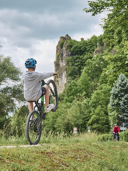 Ein Junge macht mit seinem Fahrrad einen Wheelie auf dem Radweg umringt von grüner Natur.