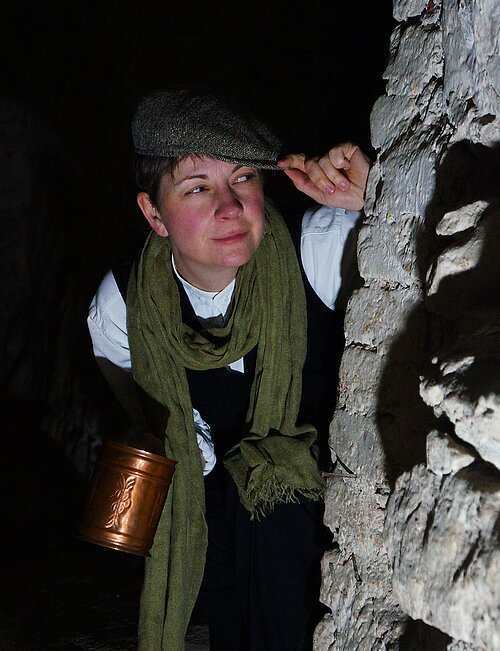 Kellergrattler Person mit Schirmmütze und grünem Schal hält Kupfergefäß und blickt an Steinmauer im Dunkeln vorbei.