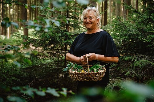 Frau mit schwarzem Oberteil hält Korb mit gesammelten Kräutern im Wald stehend.