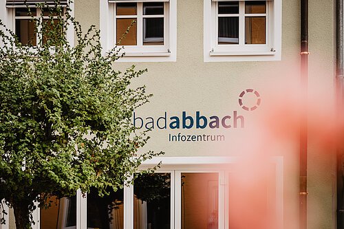 Fassade eines Gebäudes mit Schriftzug „bad abbach Infozentrum“ und zwei Fenstern, links ein Baum.