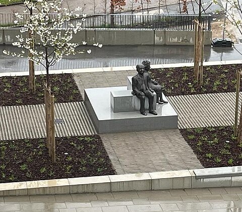 Skulptur von zwei sitzenden Personen auf einem mehrstufigen Podest in einem bepflanzten Stadtbereich mit blühenden Bäumen.