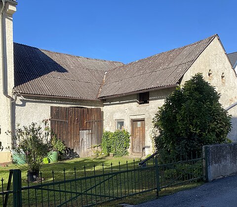 Hof mit altem Gebäude, Holztüren, grünem Metallzaun, Büschen und blauem Himmel im Hintergrund.