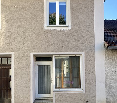 Fassade eines Hauses mit weißem Eingangstür, großem Fenster und kleinem Fenster darüber, Pflastersteine davor.
