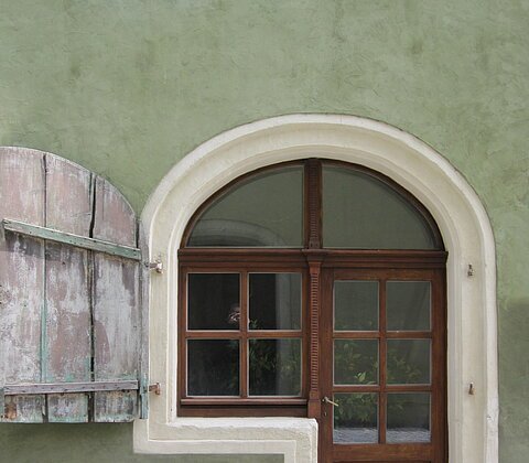 Holztür mit Fenster im Bogen und geöffnetem Fensterladen an grünem Putz mit Kopfsteinpflaster davor.