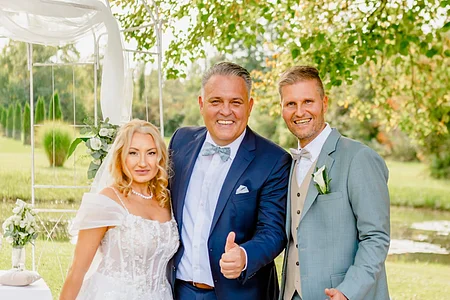 Braut in weißem Kleid, Mann in blauem Anzug und Mann in grauem Anzug mit Daumen hoch im Garten bei Hochzeit