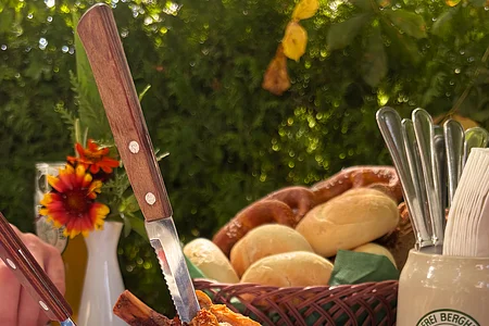 Teller mit Schweinshaxe, Krautsalat und Zwiebeln auf blau-weiß kariertem Tisch mit Brötchenkorb im Hintergrund.