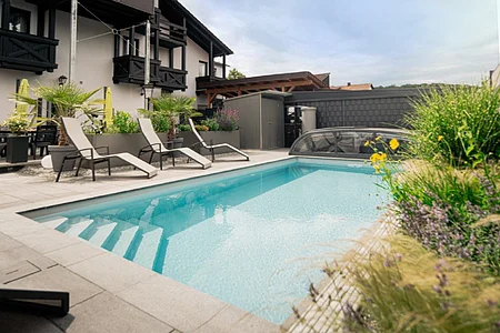 Außenbereich mit rechteckigem Pool, umgeben von Liegestühlen, Pflanzen und einem Gebäude im Hintergrund.