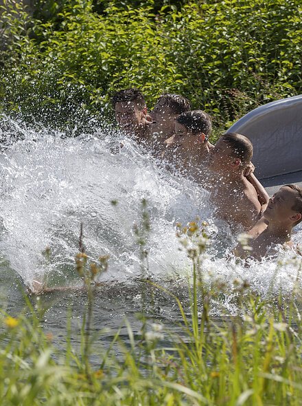 Fünf Jungen rutschen gemeinsam eine Wasserrutsche hinunter und spritzen ins Wasser, umgeben von grüner Vegetation.