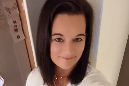 Eine Person mit dunklem Haar macht ein Selfie in einem Raum mit Holzboden und Kühlschrank im Hintergrund.