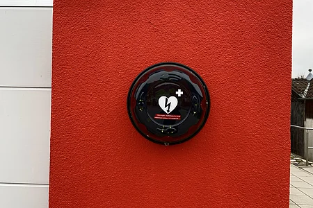 Defibrillator in schwarzem Gehäuse an roter Wand montiert, links weiße Wandpaneele sichtbar.