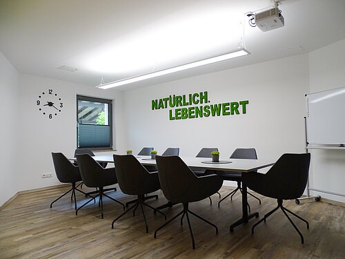 Konferenzraum mit Tisch, acht Stühlen, Uhr, Fenster, Whiteboard und Schriftzug „Natürlich. Lebenswert“ an der Wand.