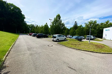 Parkplatz mit mehreren Autos, umgeben von Bäumen und einem Gebäude mit Schriftzug „Asklepios“