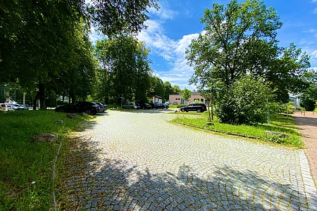 Parkplatz mit mehreren Autos, umgeben von Bäumen und Gebäuden unter blauem Himmel mit Wolken