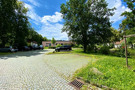 Parkanlage mit gepflastertem Parkplatz, mehreren geparkten Autos, Bäumen und Gebäuden im Hintergrund bei sonnigem Himmel.