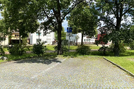 Leerer gepflasterter Parkplatz mit Schild für Behindertenparkplatz und Bäumen im Hintergrund.