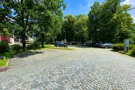 Parkplatz mit Kopfsteinpflaster, mehreren geparkten Autos und Bäumen bei sonnigem Himmel