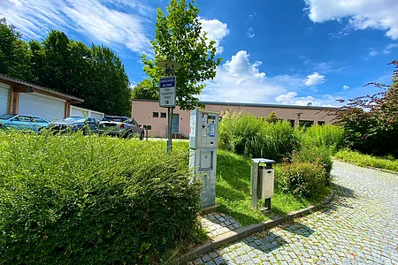 Parkautomat und Mülleimer an gepflastertem Weg mit parkenden Autos und Gebäuden im Hintergrund bei sonnigem Himmel.