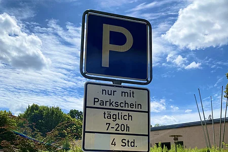 Blauer Parkplatzschild mit Zusatzschildern „nur mit Parkschein täglich 7-20h 4 Std. Parken in gekennzeichneten Flächen erlaubt“ vor grüner Bepflanzung.
